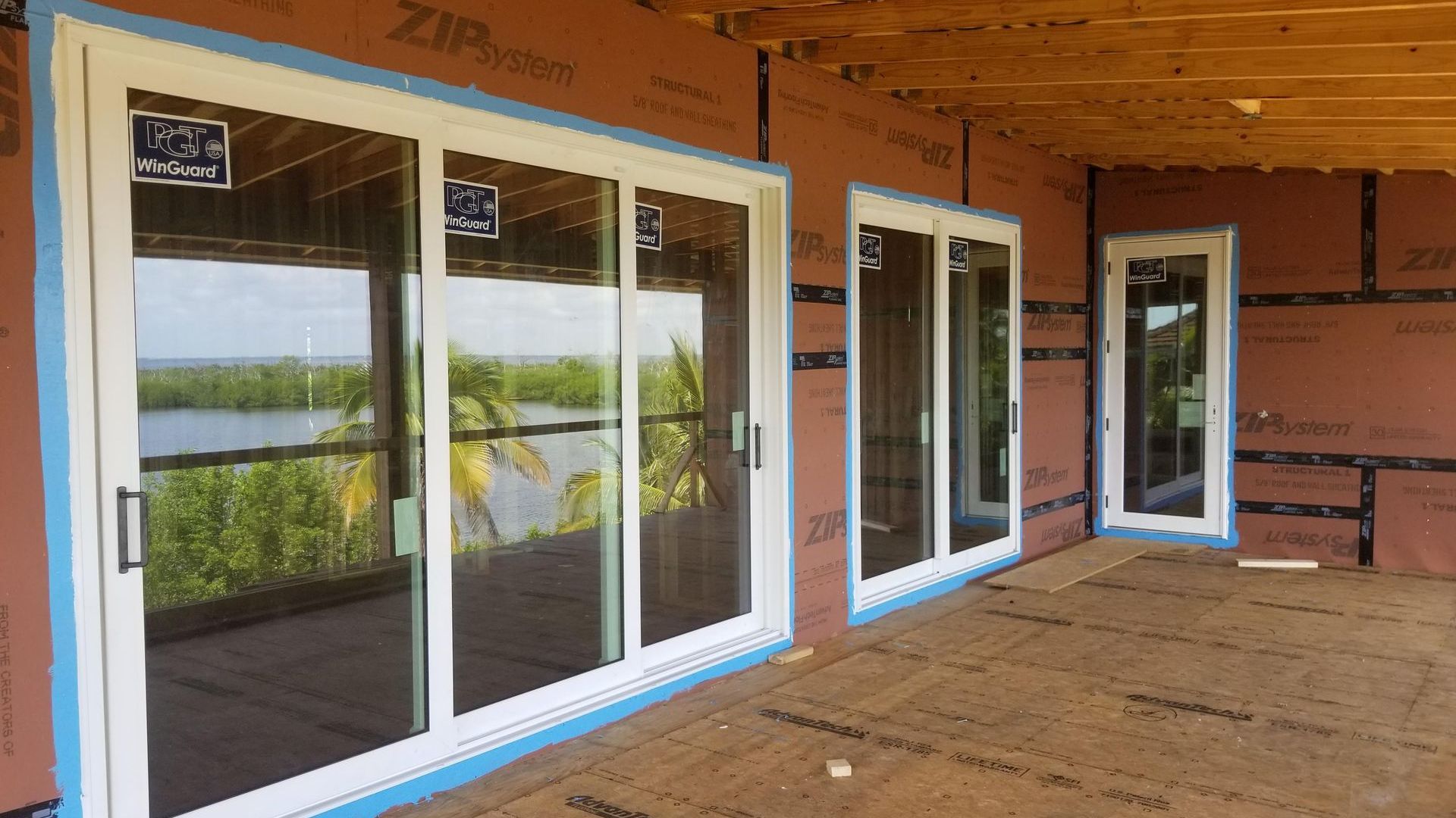 Exterior view of a house under construction with three white-framed glass sliding doors. The background shows water and trees.