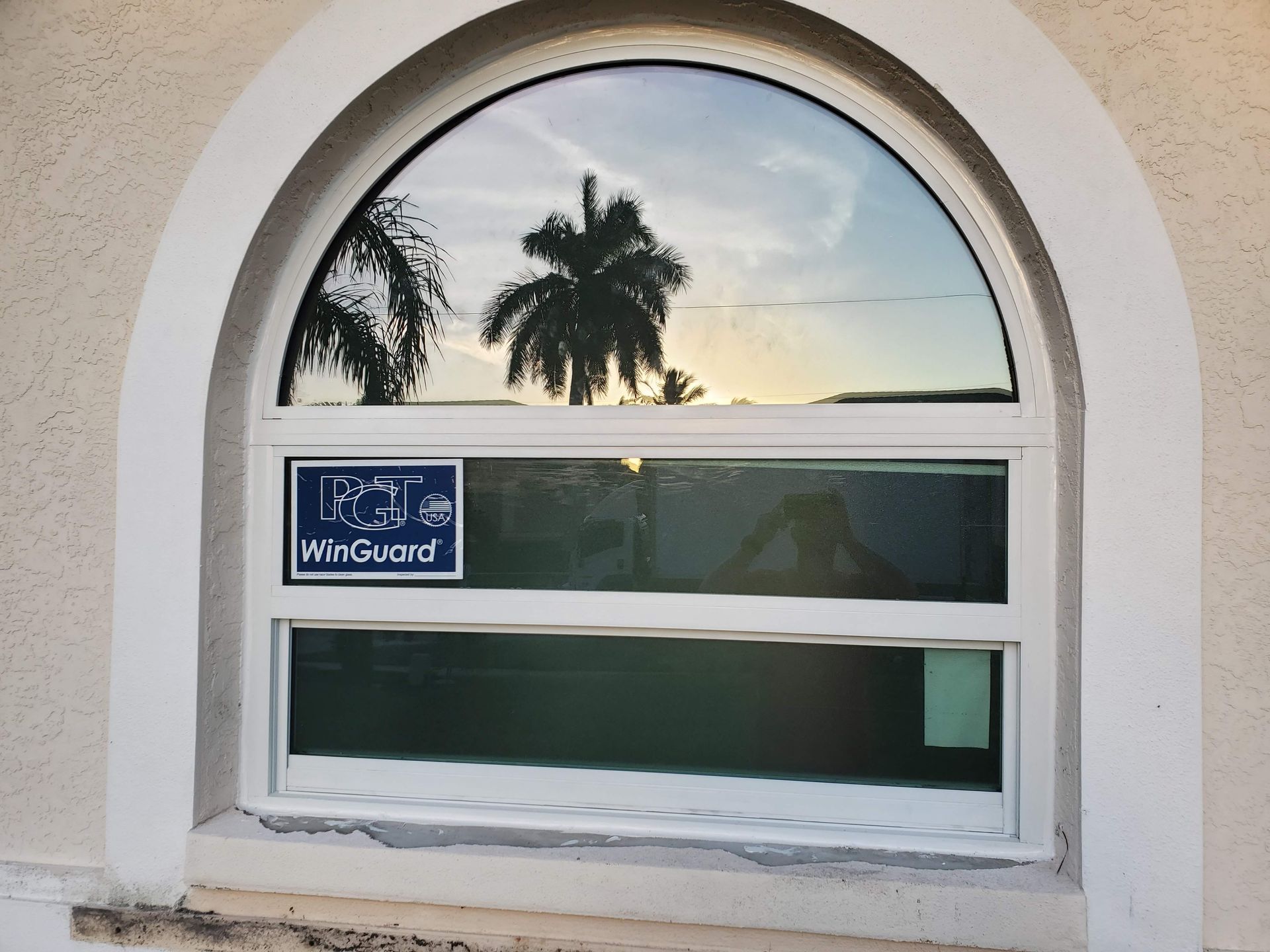 Arched window with a white frame, reflecting trees and sky, with a WinGuard sticker.