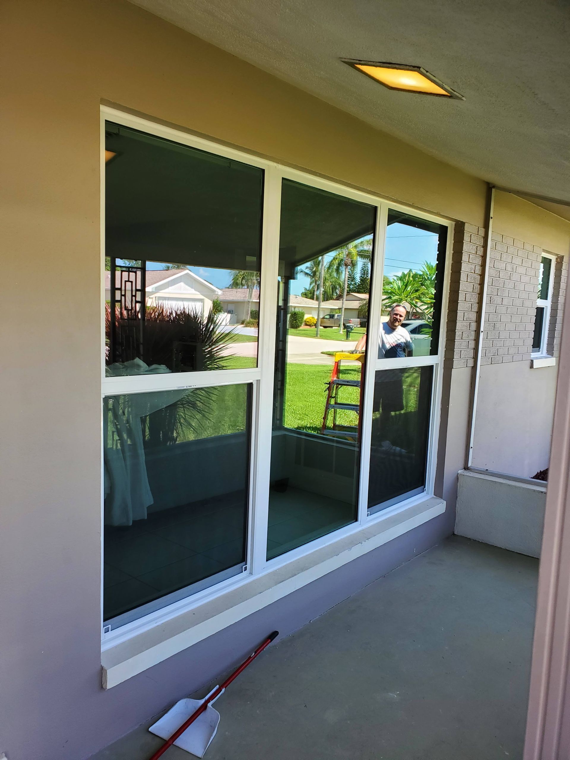 A large window with white trim reflects a green yard and blue sky. A broom rests on the porch.