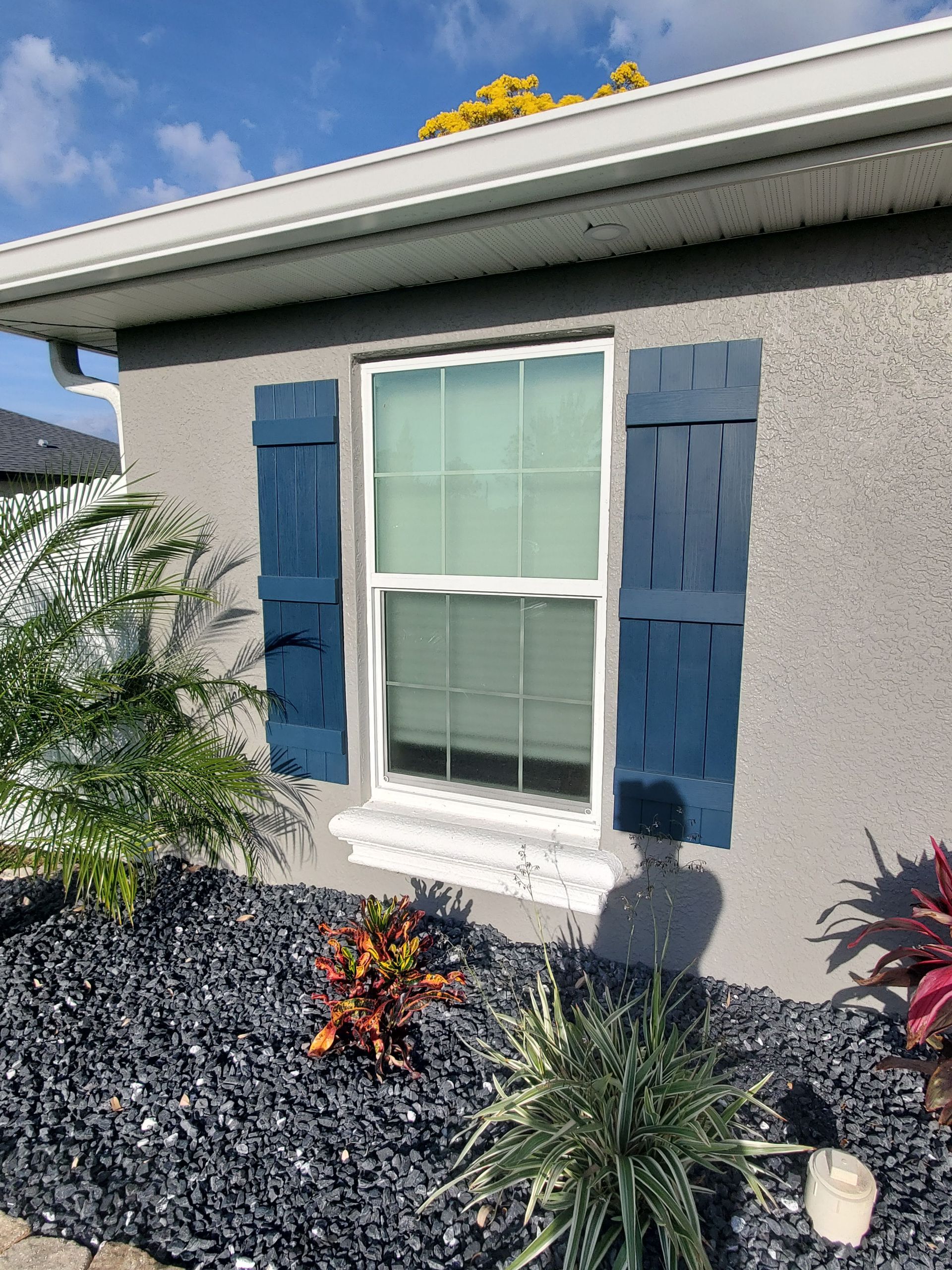Window with blue shutters on a gray stucco building, surrounded by dark rocks and plants.