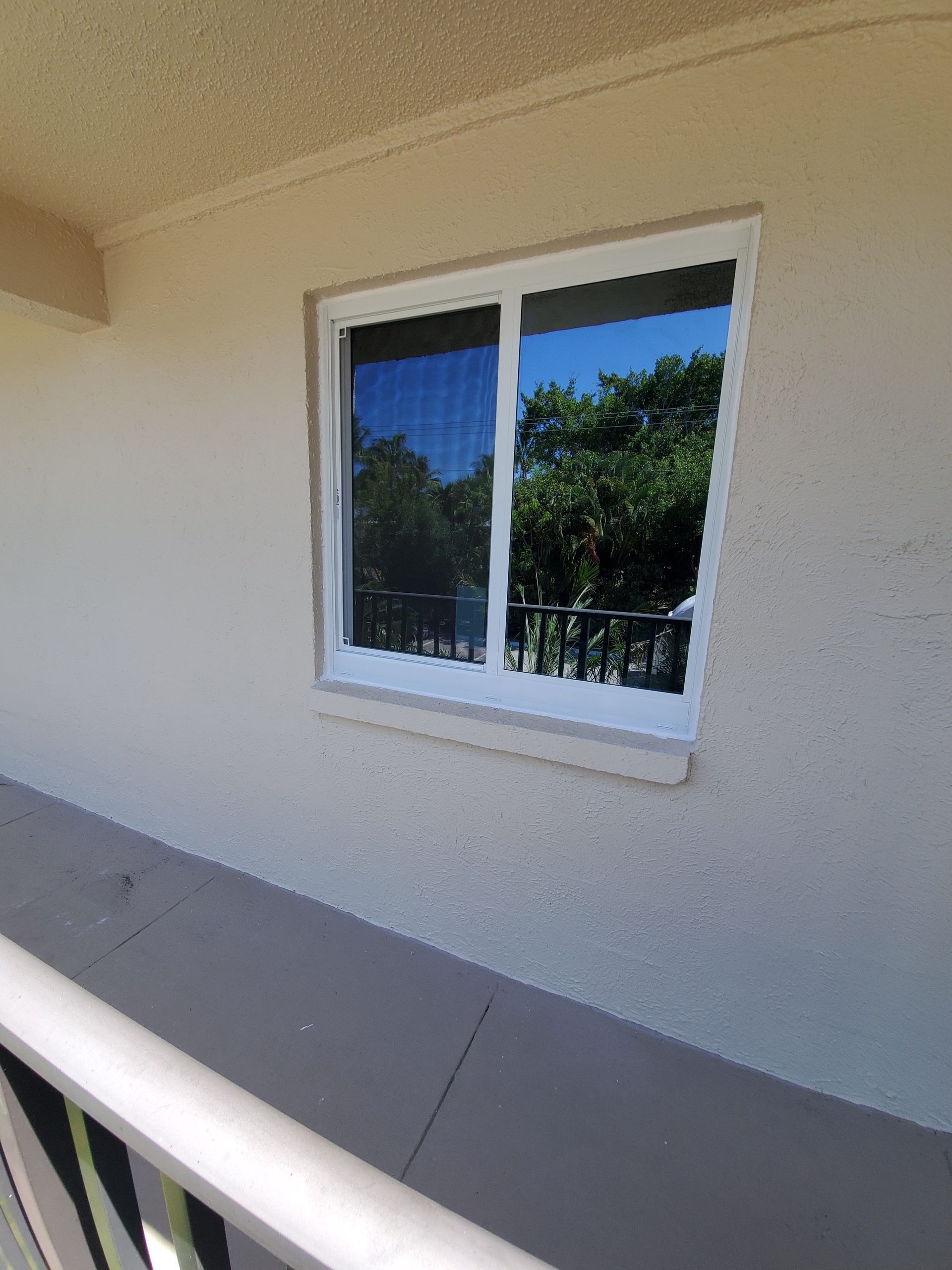 Window on a beige building, reflecting trees and sky. A balcony railing runs along the bottom.