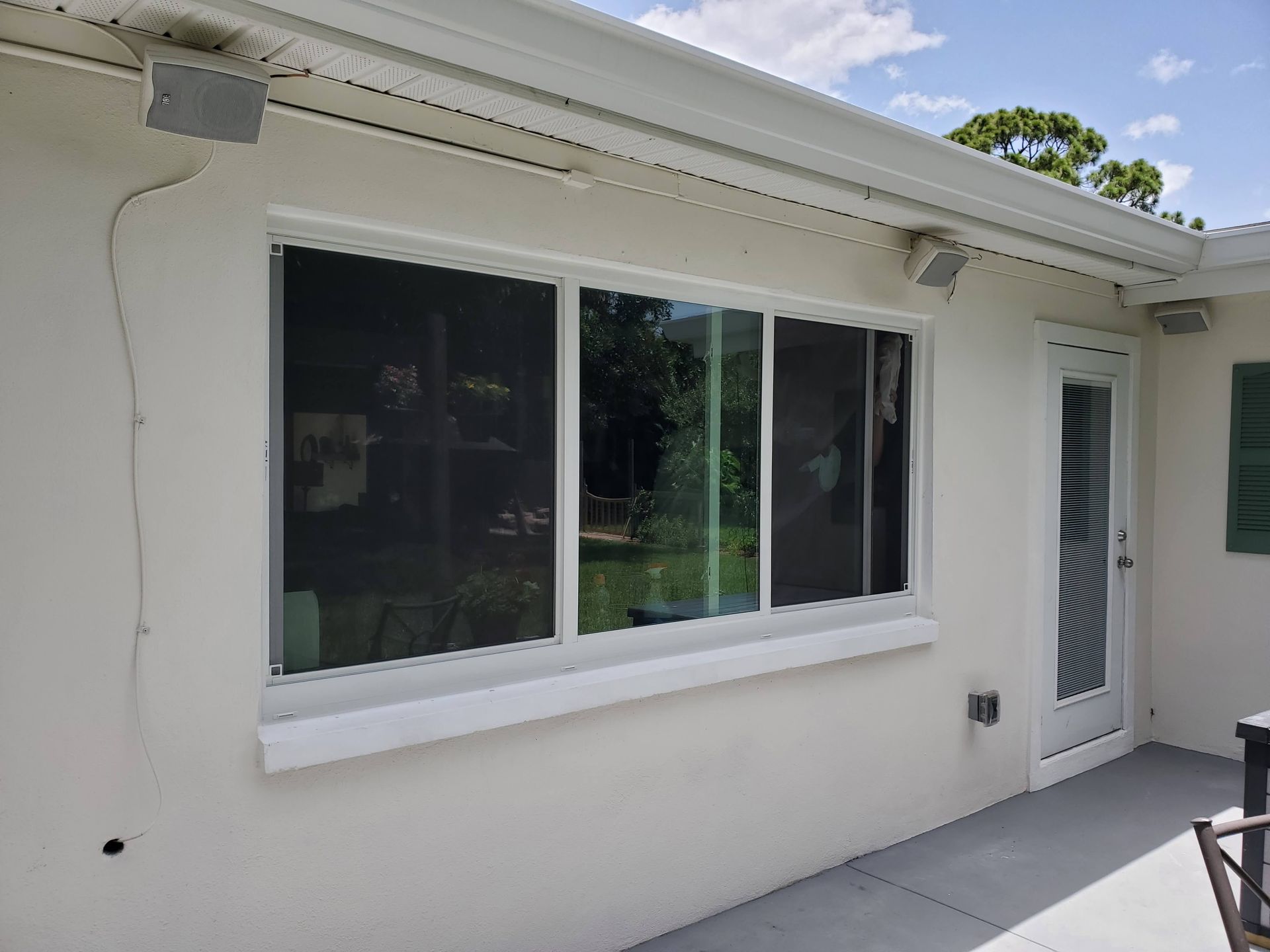 Exterior view of a white stucco wall with a sliding glass window and door, speakers mounted above.