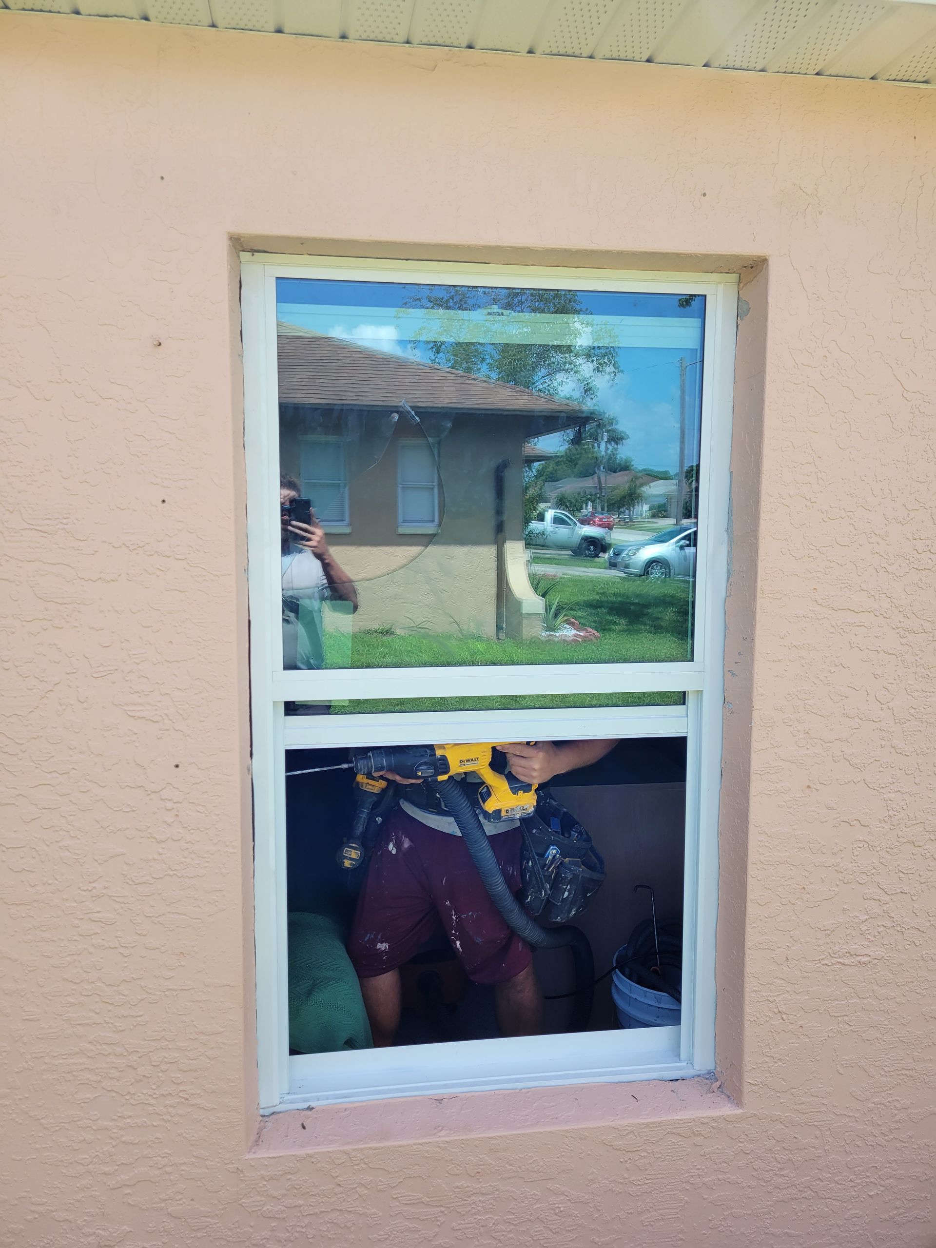 A person installs a window. They use a drill while working. The house exterior is peach-colored.