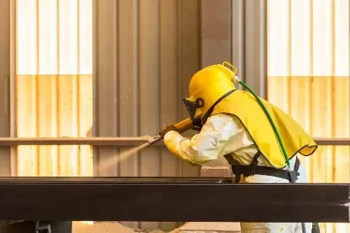 A Man Wearing A Yellow Helmet Is Sandblasting A Piece Of Metal — Rockhampton Powdercoating & Fence Panels in Park Avenue, QLD