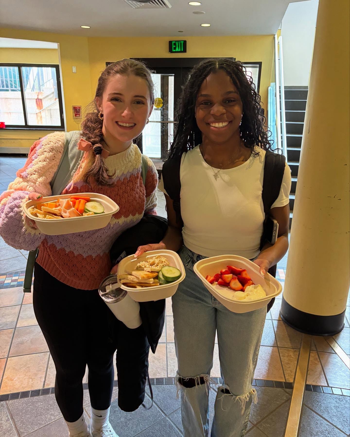 Two women are standing next to each other holding plates of food.