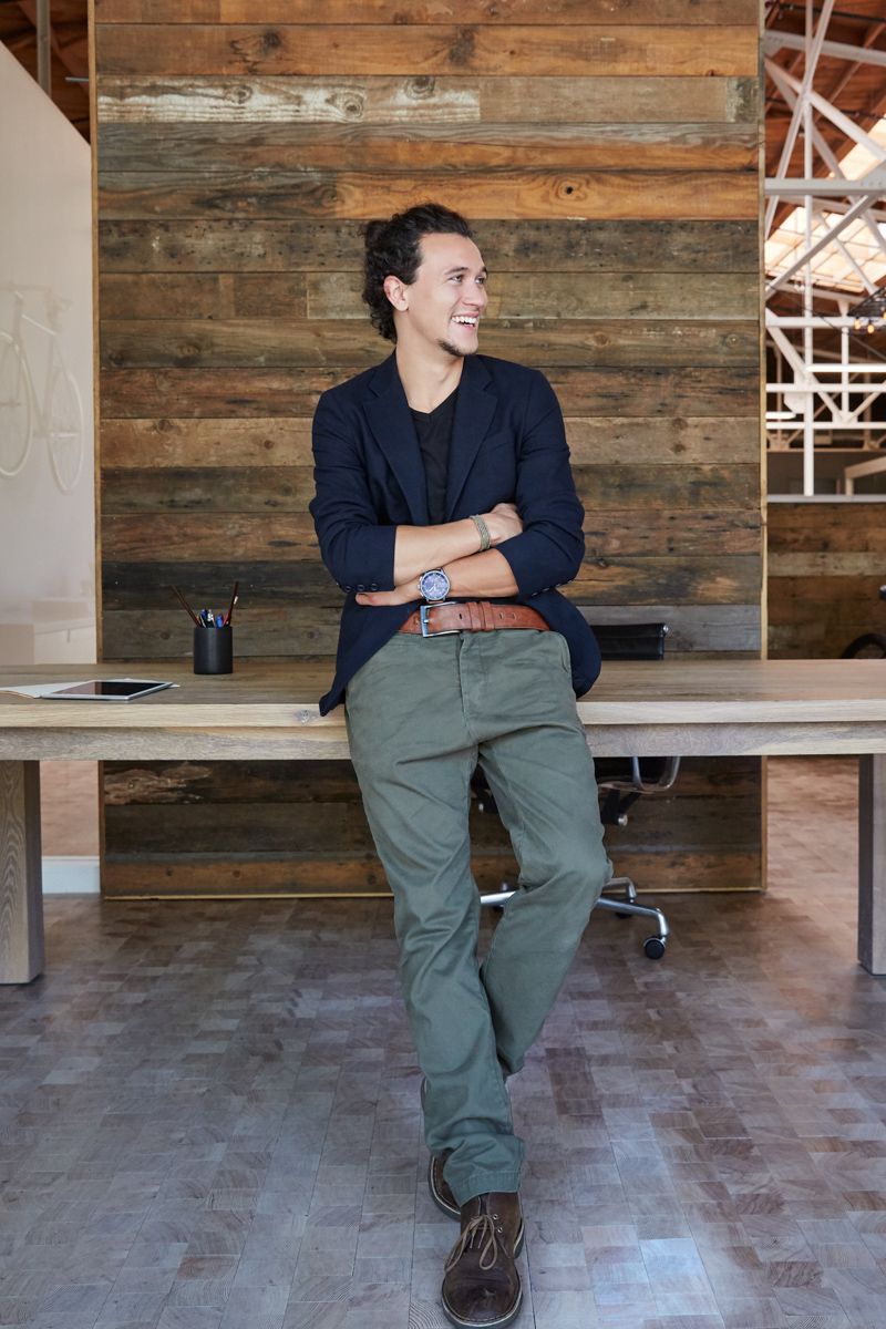 A person in a dark blazer and olive pants smiles, leaning against a wooden table in a rustic, modern office space.