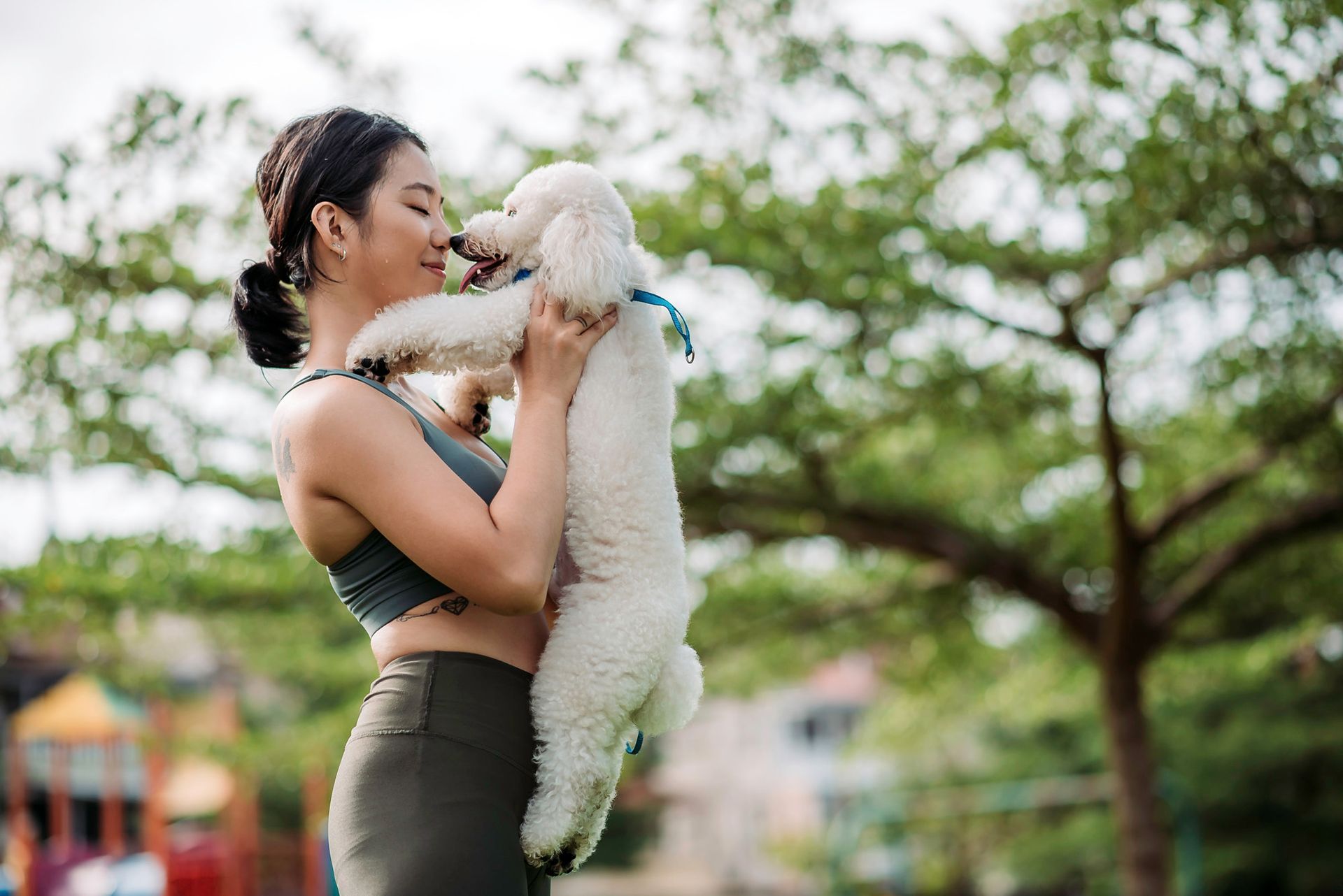 A person in athletic wear stands outdoors holding a fluffy white dog up toward their face in an affectionate embrace.