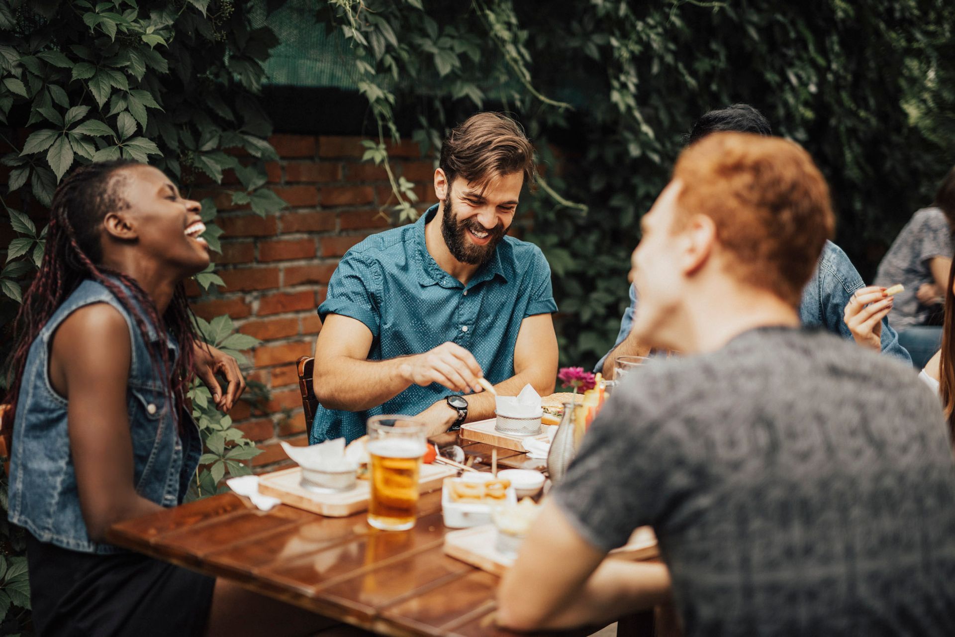 Friends laughing and eating together at an outdoor wooden table with drinks, set against a leafy brick wall.