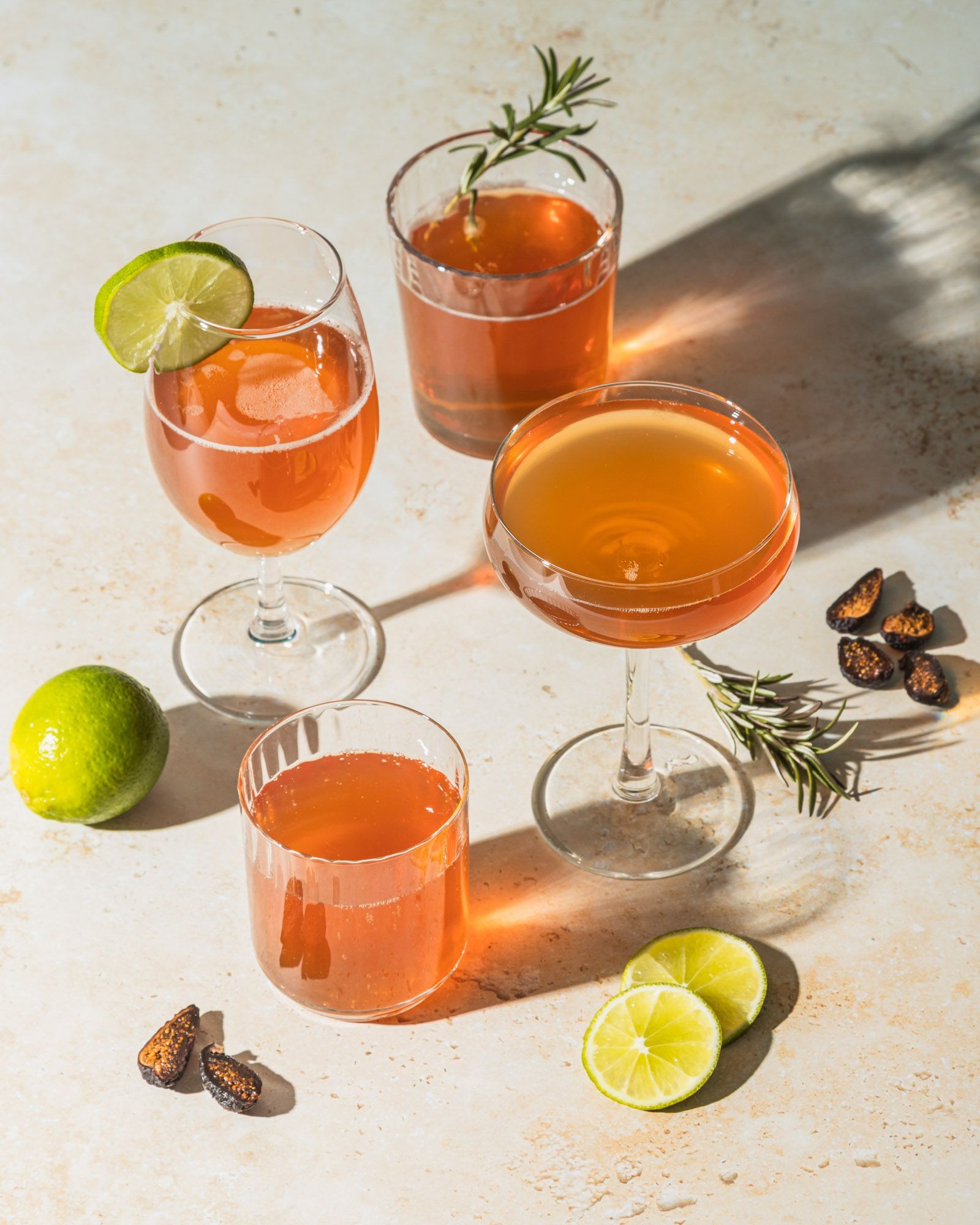 Four amber-colored cocktails in various glasses, garnished with rosemary and lime slices on a textured light surface.