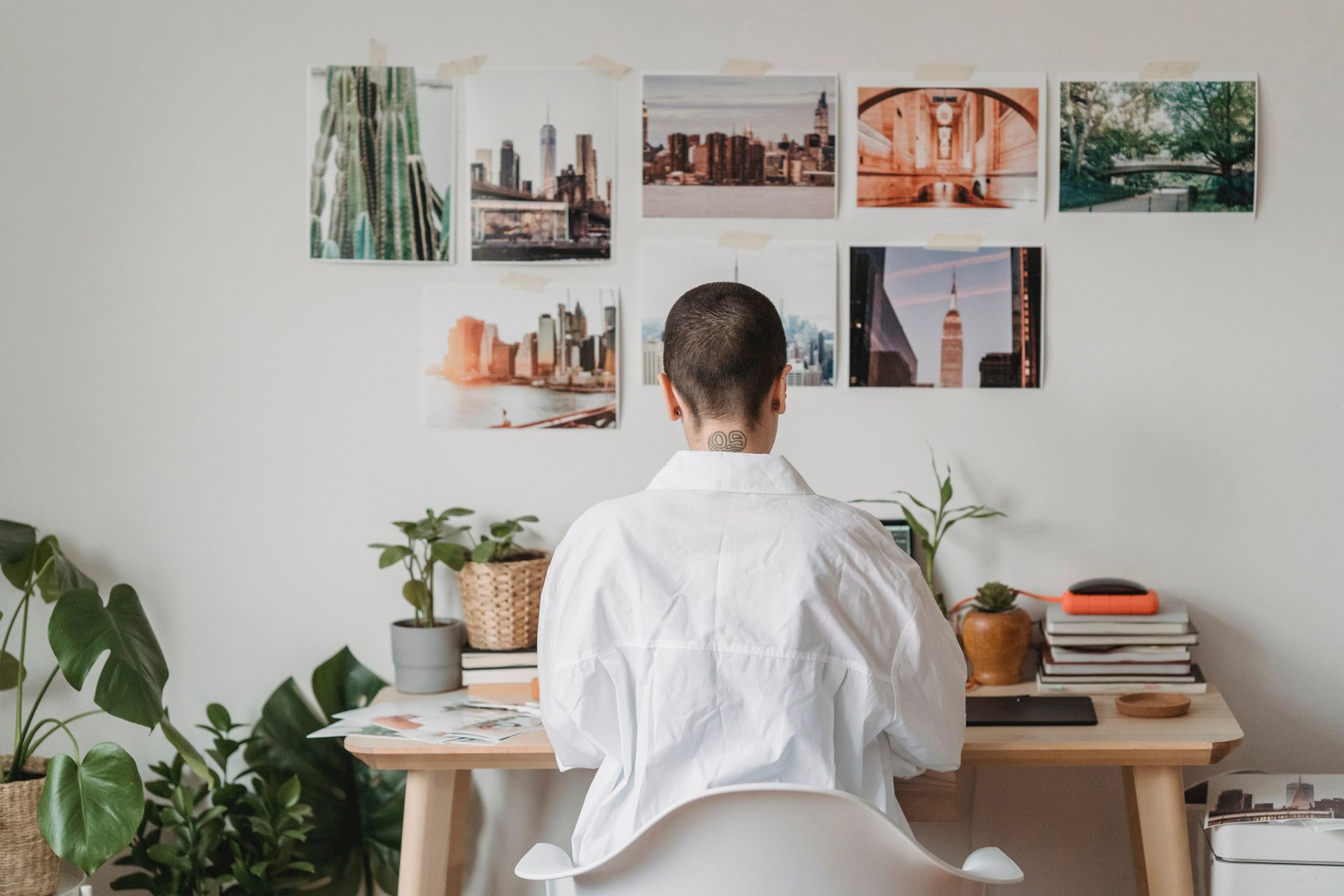 A person in a white shirt works at a wooden desk with plants, facing a white wall covered in assorted framed city photos.