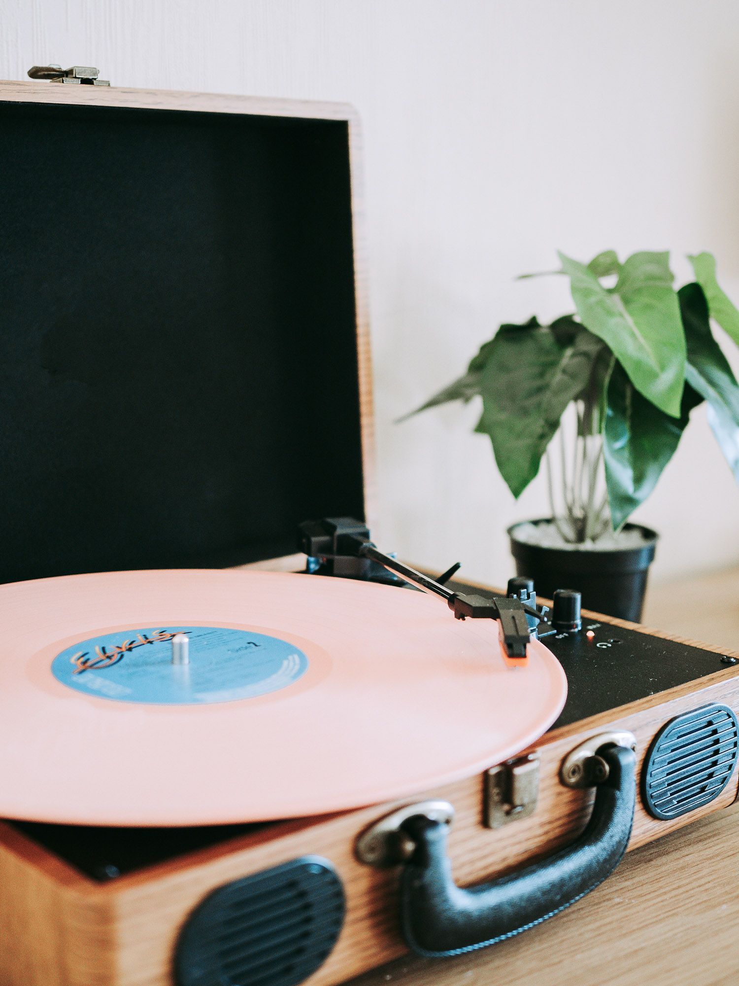 A pink vinyl record plays on a portable, wood-paneled record player next to a potted green plant.