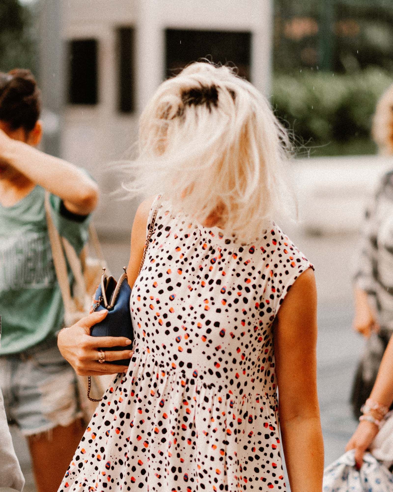 A person with windswept blonde hair wears a sleeveless, white spotted dress while carrying a small blue handbag.