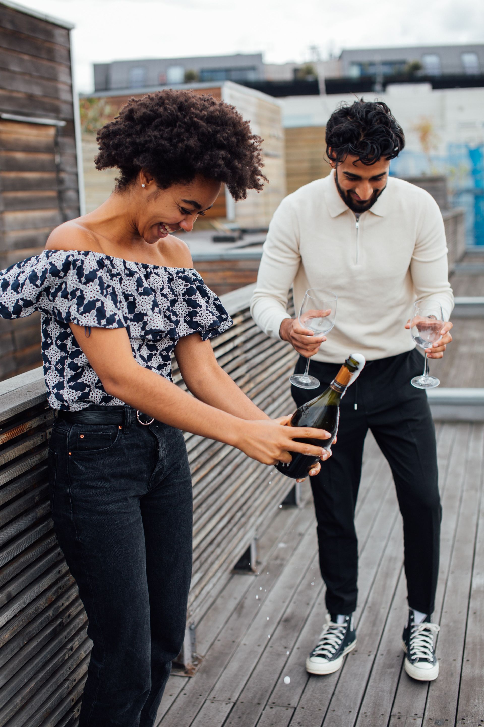 A smiling person opening a bottle of champagne on a wooden rooftop deck, while another person holds two empty glasses.