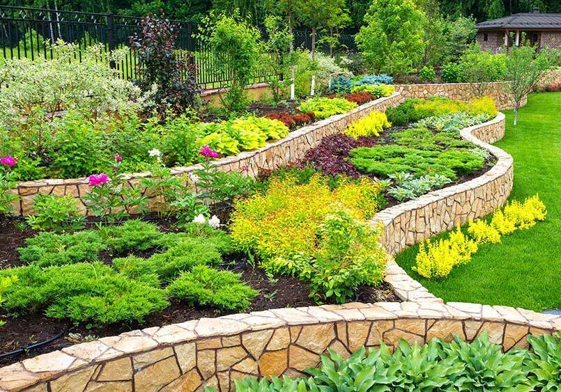 A garden with lots of plants and flowers and a stone wall.