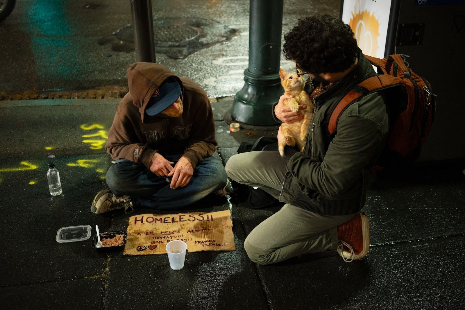 Two people on a wet sidewalk with a cat, one kneeling holding the cat, the other seated by a