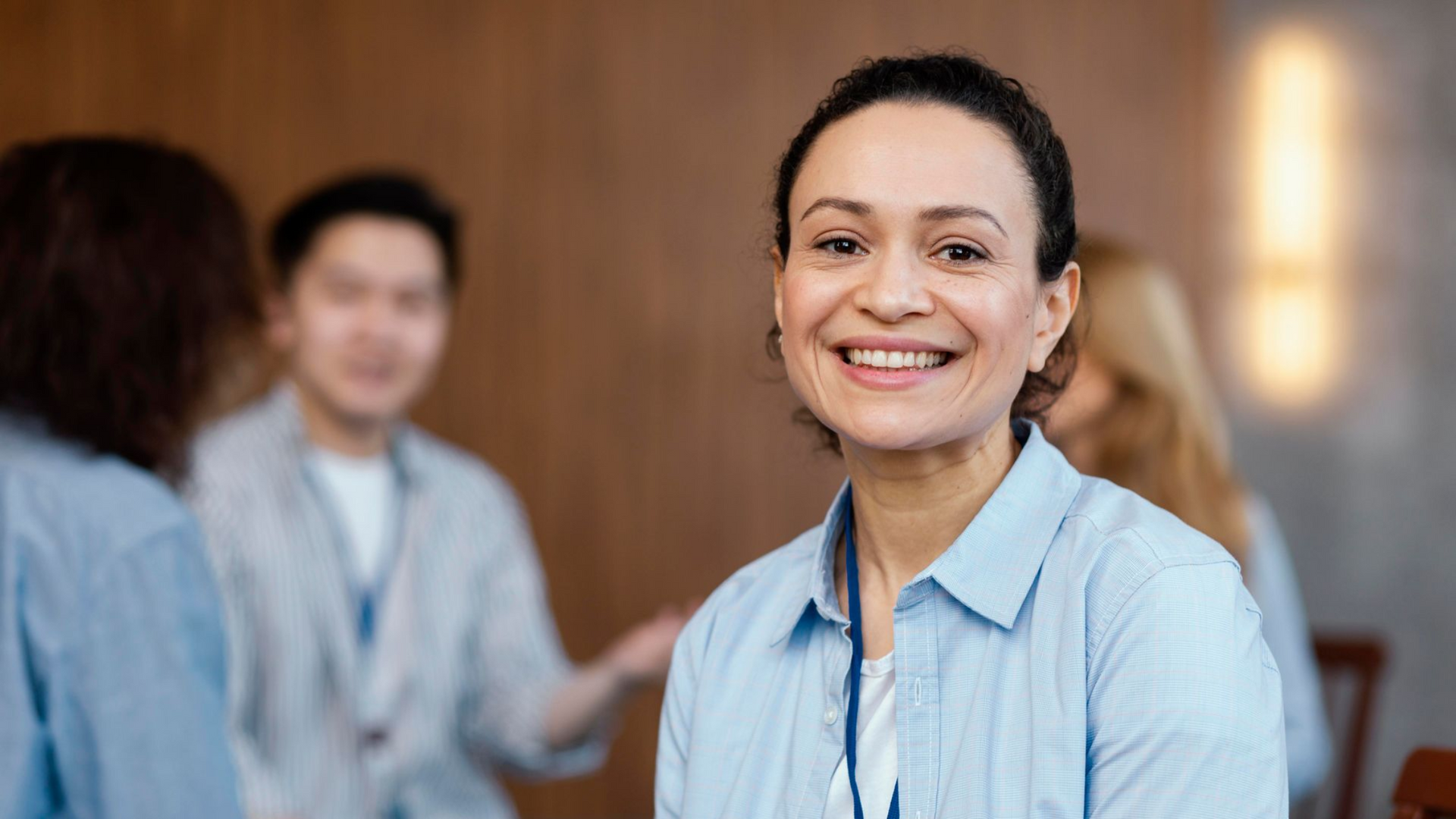 Woman smiling at camera, sitting in a room with others. She wears a blue button-down shirt.