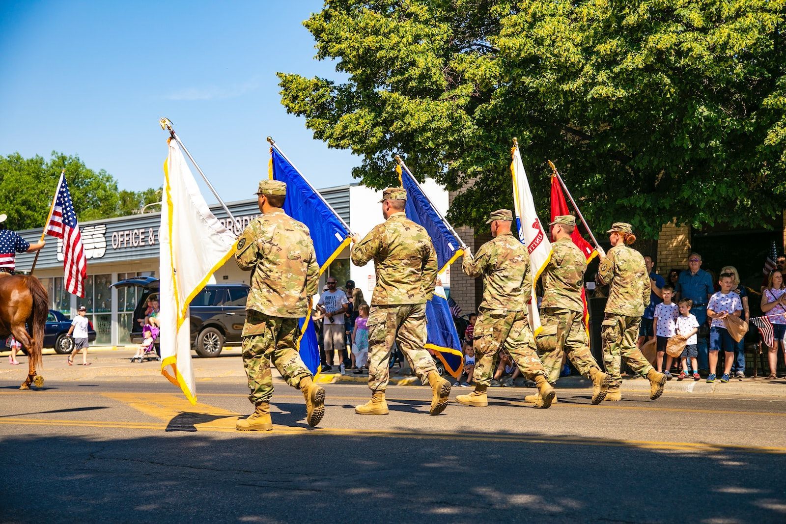 Soldiers in camouflage uniforms carry flags during a parade on a sunny day.