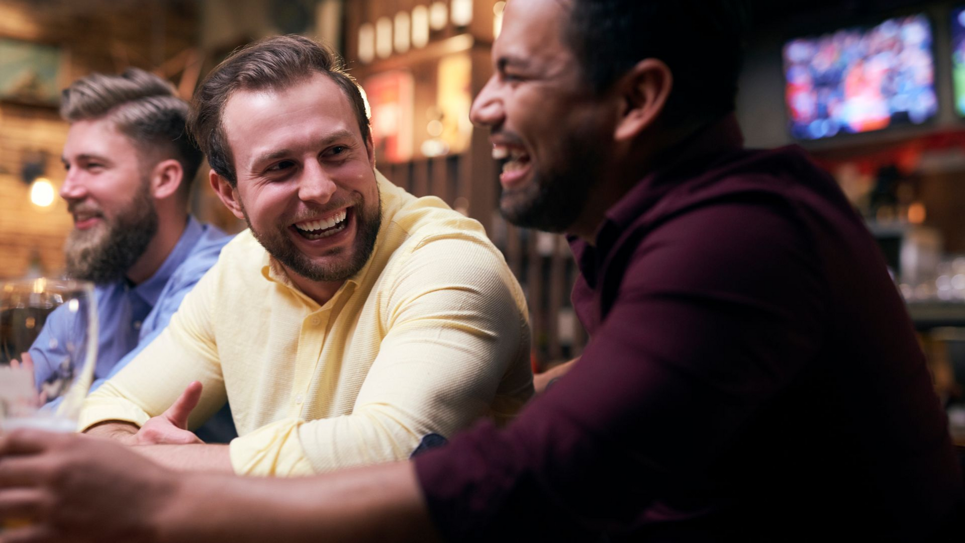 Three men laugh while sitting at a bar. One wears a yellow shirt, another a burgundy shirt. Dark bar setting.