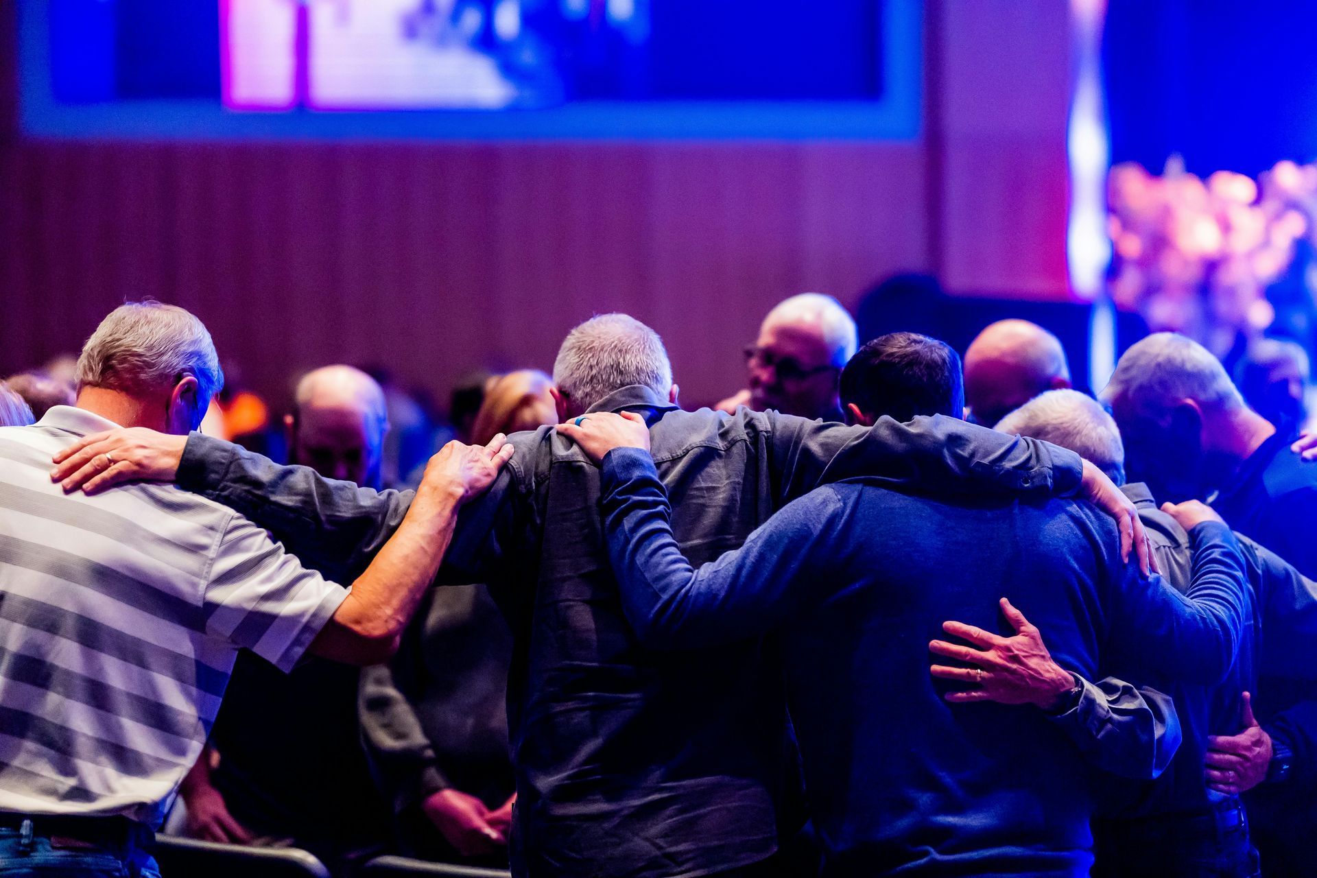 Group of men with arms around each other, praying together. Purple and blue lighting, indoor setting.
