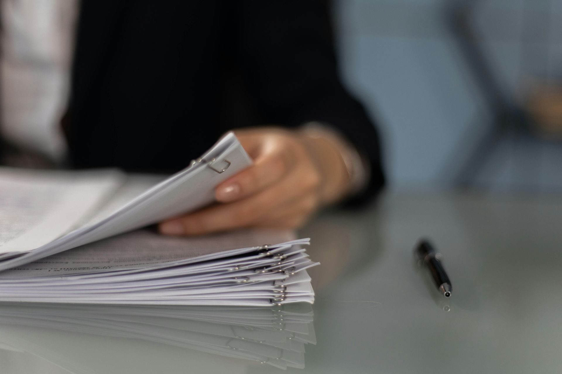 Person's hand sorting through a stack of papers, a pen on the desk.