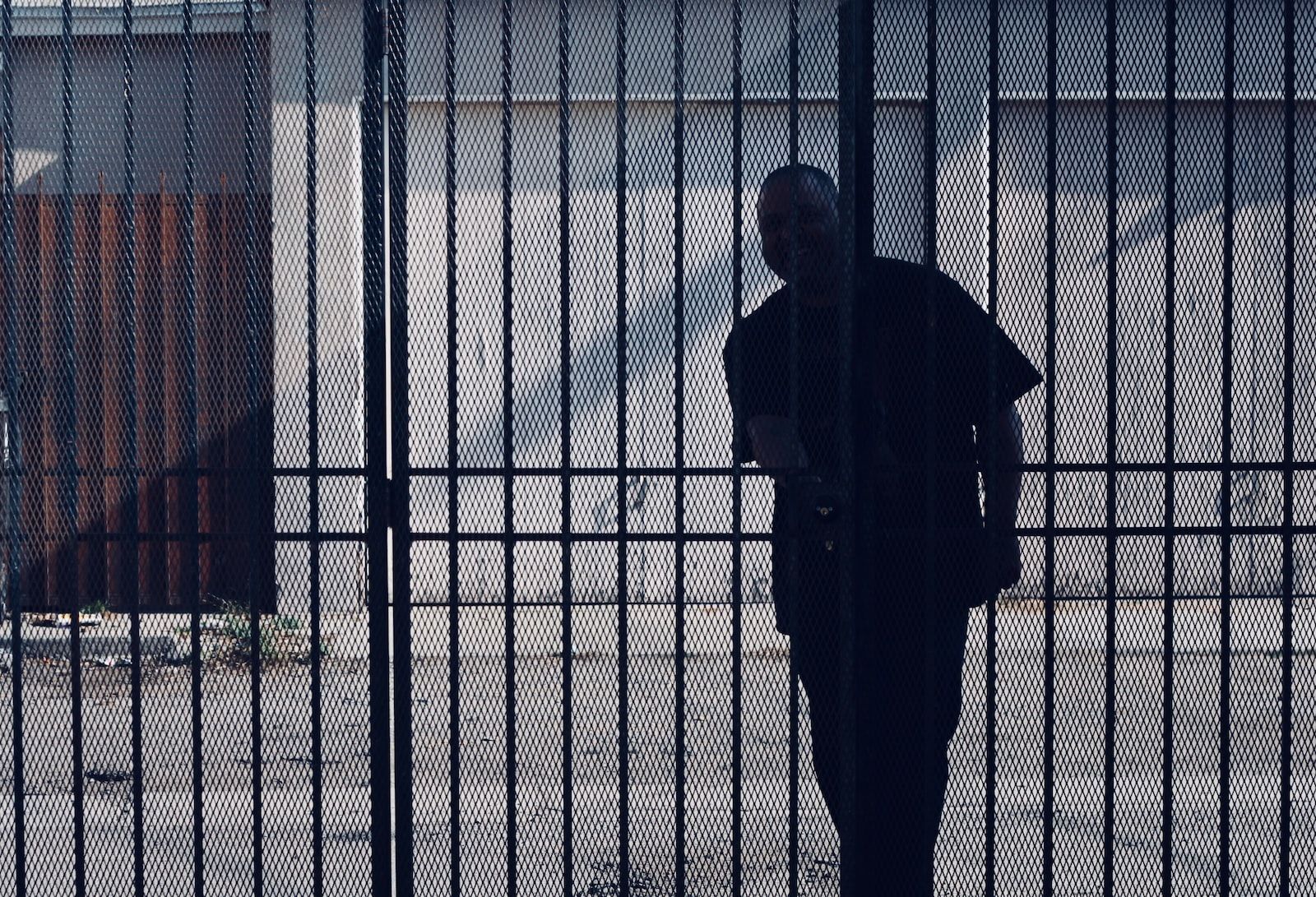 Man silhouetted behind a metal gate, standing near a building with a rust-colored section.