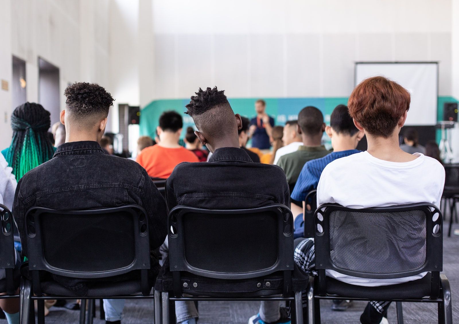 Audience seated in a large room, facing a speaker near a screen. Black mesh chairs.