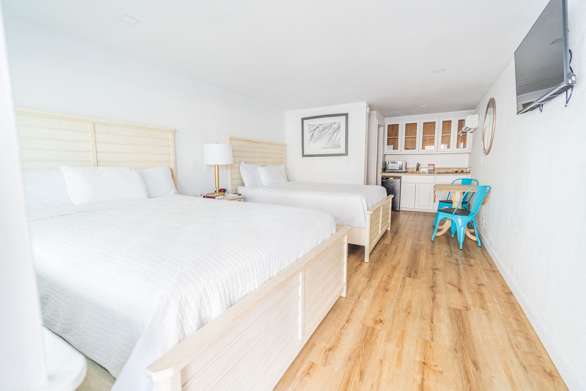 Bright hotel room with two beds, small kitchenette, and wooden floors.
