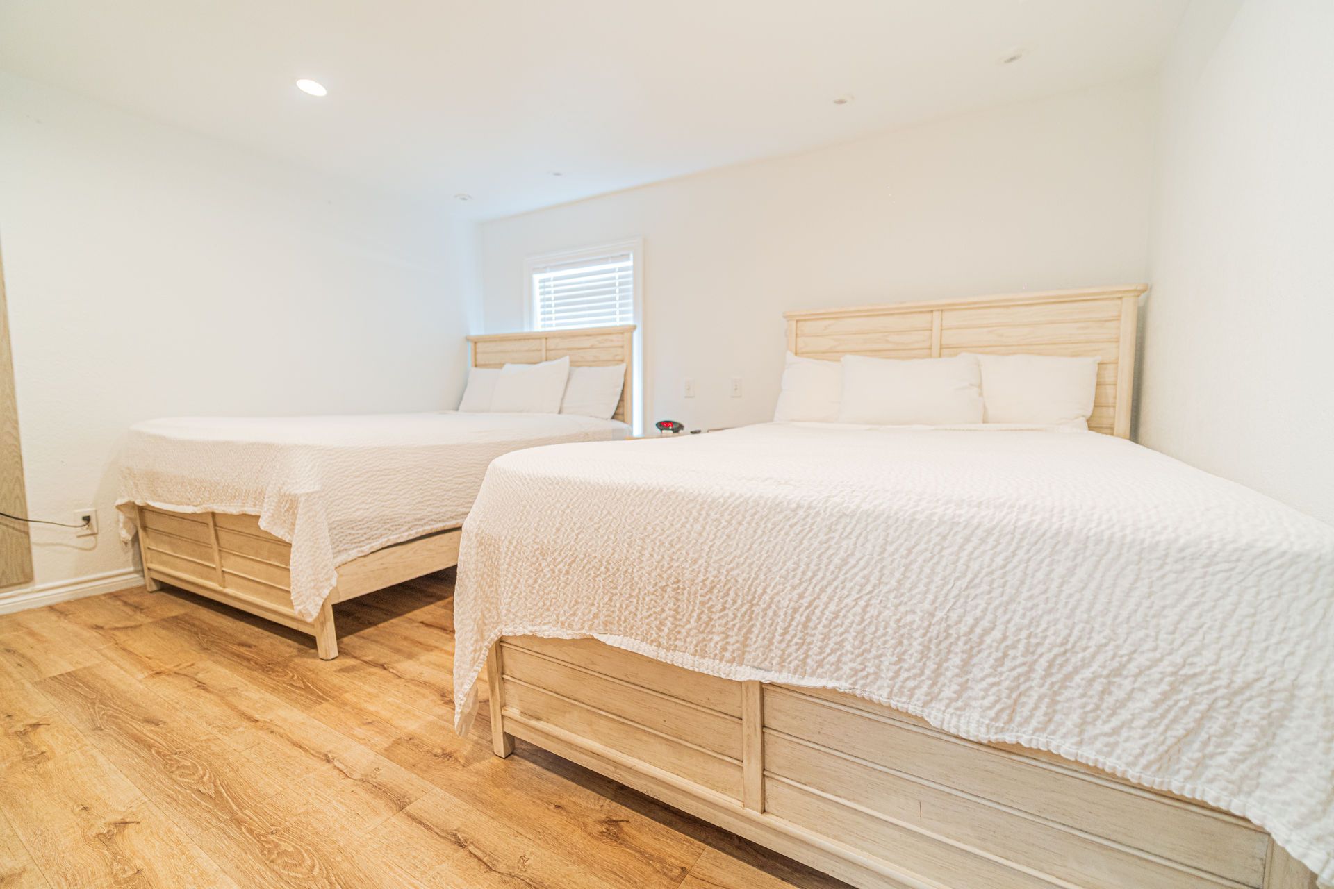 Two beds in a white-walled room with light wood floors. Each bed has a white quilt and headboard.