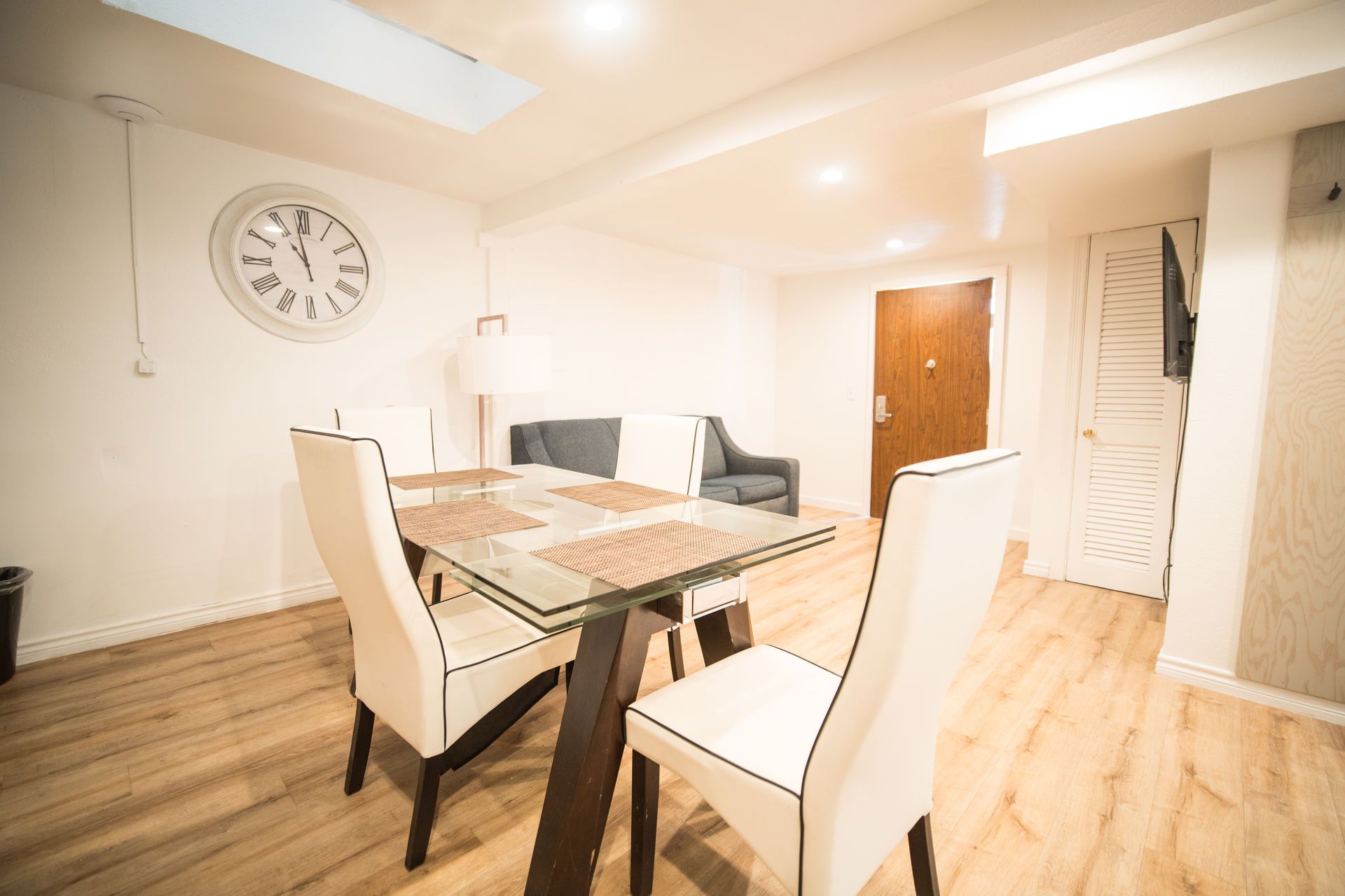 Dining area with a glass table, white chairs, a clock, and a sofa.