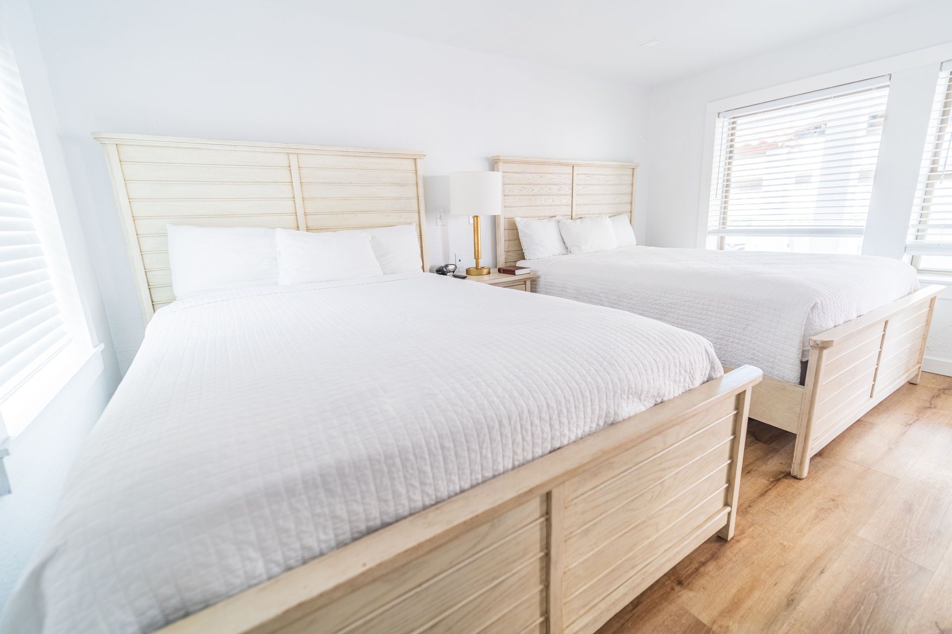 Two beds in a bright room with white bedding and wooden frames, near windows with blinds.