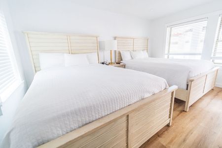 Two beds in a bright room with white bedding and wooden frames, near windows with blinds.