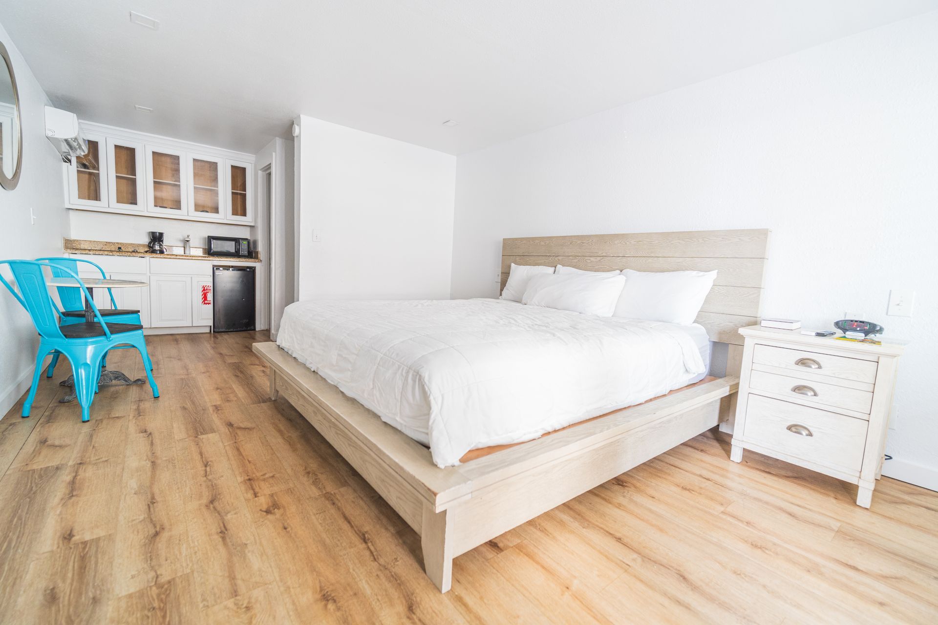 Bedroom with light wood flooring, bed, small kitchen area, blue chair.