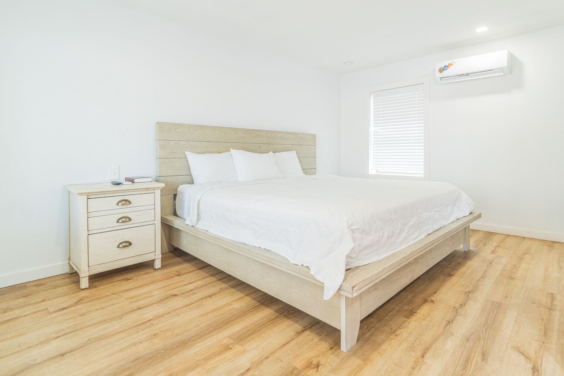 Bedroom with bed, nightstand, and air conditioner on a light wood floor. White walls.