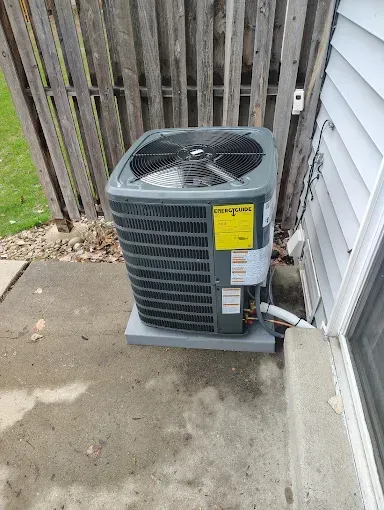 Air conditioner unit outside, next to a building and fence, on a concrete pad.