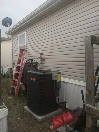 Exterior view of a house with an air conditioning unit. Red ladder, brown siding. Overcast day.