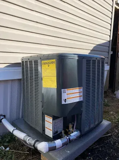 An air conditioning unit outdoors, gray metal, yellow energy label, connected to white pipes, near siding.