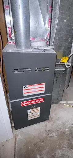 Goodman furnace in a basement. The furnace is gray and has red labels. There is a metal vent pipe.