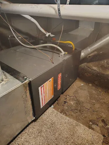 Furnace in a basement, gray metal with orange and red stickers, connected to ductwork and gas line.