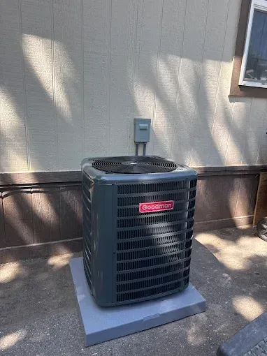 Gray Goodman air conditioning unit on a concrete pad next to a wall with electrical box.