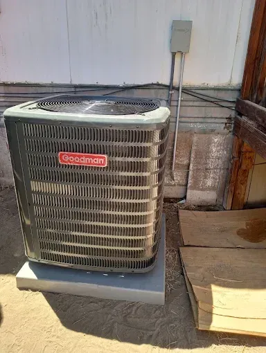 Goodman air conditioning unit on a concrete pad next to a wall. Gray and beige.