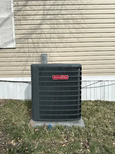 Gray Goodman air conditioner on concrete base next to a tan house with a vent pipe.