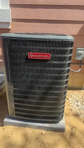 Goodman air conditioning unit, outdoor setting, black metal grill, red logo, gray concrete base.