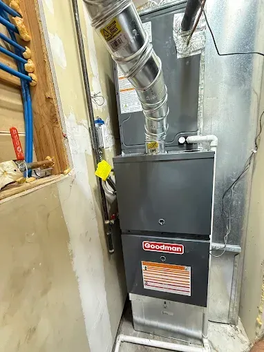 A new Goodman furnace installed in a narrow, unfinished utility room. Silver ductwork is connected.