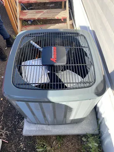 Air conditioner unit outside, gray, with a black fan cage and red emblem on top.