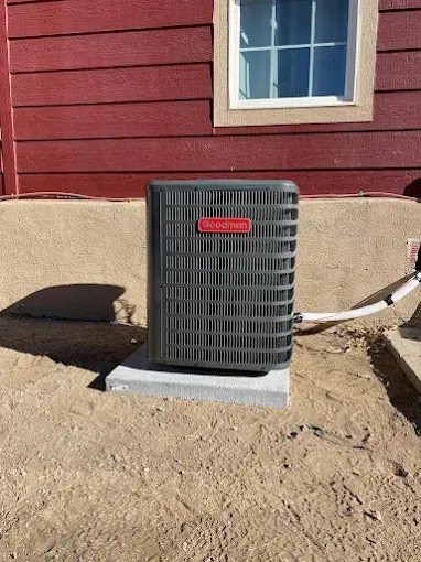An air conditioning unit on a concrete block next to a red-sided building, with a window visible.