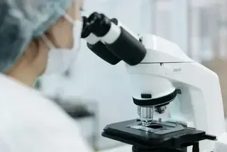Scientist wearing a mask and cap using a white microscope in a lab setting.
