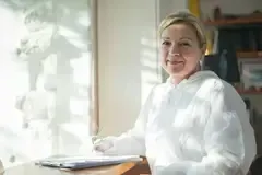 Woman in white lab coat smiles while writing at a desk, with sunlight and a window behind her.