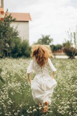 Woman with red hair runs through a field of white flowers, wearing a white dress.