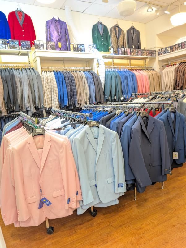 Rows of men's suit jackets in various colors, displayed on rolling racks in a retail store.