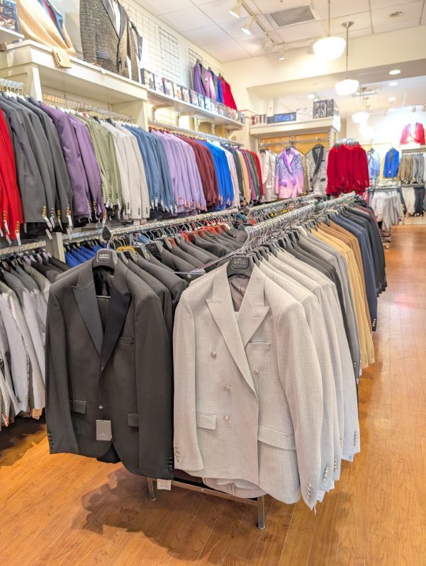 Rows of colorful suit jackets hang in a well-lit clothing store with wooden floors and white walls.